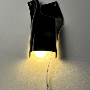 May include: A wall-mounted lamp featuring a glossy black, curved, sculptural shade that directs light downward. A single warm-toned light bulb is visible within the opening, and a clear electrical cord hangs vertically from the bottom of the fixture.