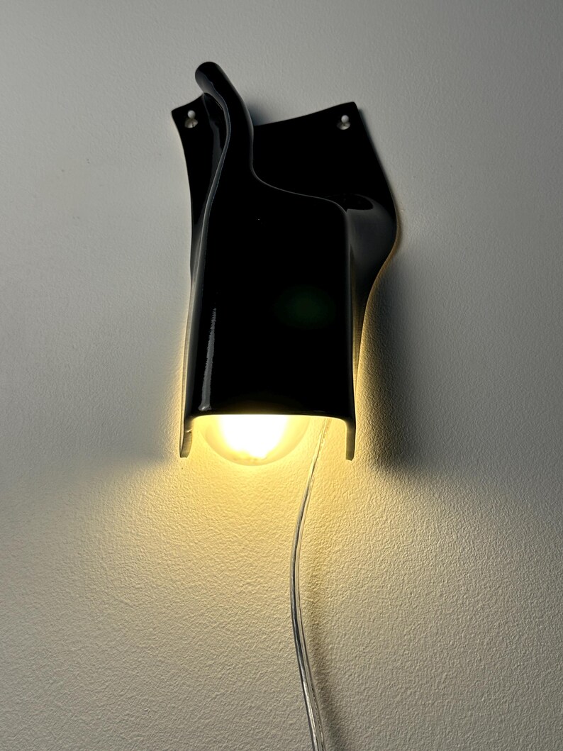May include: A wall-mounted lamp featuring a glossy black, curved, sculptural shade that directs light downward. A single warm-toned light bulb is visible within the opening, and a clear electrical cord hangs vertically from the bottom of the fixture.