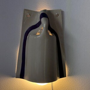 May include: A wall-mounted ceramic sconce featuring a glossy off-white finish with a dark purple vertical stripe accent. The fixture has a curved, sculptural form that emits a warm, soft glow from both the top and bottom openings.