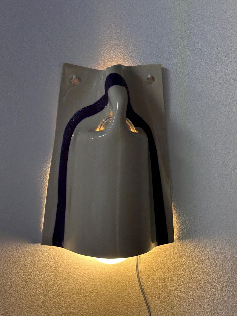 May include: A wall-mounted ceramic sconce featuring a glossy off-white finish with a dark purple vertical stripe accent. The fixture has a curved, sculptural form that emits a warm, soft glow from both the top and bottom openings.