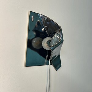 Peut inclure: Une applique murale dot&eacute;e d'un abat-jour en c&eacute;ramique bleu sarcelle fonc&eacute; brillant, pr&eacute;sentant une forme asym&eacute;trique repli&eacute;e. Le luminaire comprend une ampoule blanche ronde plac&eacute;e dans la structure en c&eacute;ramique et un cordon &eacute;lectrique transparent.