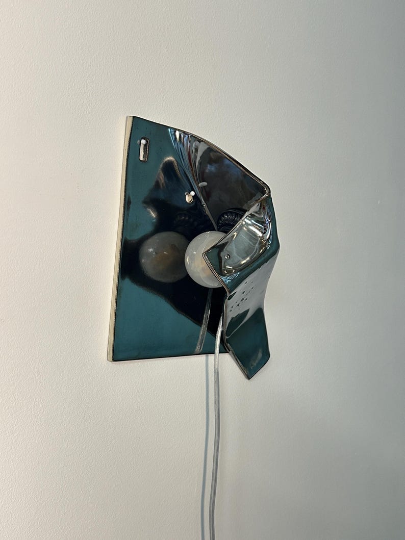 Peut inclure: Une applique murale dot&eacute;e d'un abat-jour en c&eacute;ramique bleu sarcelle fonc&eacute; brillant, pr&eacute;sentant une forme asym&eacute;trique repli&eacute;e. Le luminaire comprend une ampoule blanche ronde plac&eacute;e dans la structure en c&eacute;ramique et un cordon &eacute;lectrique transparent.