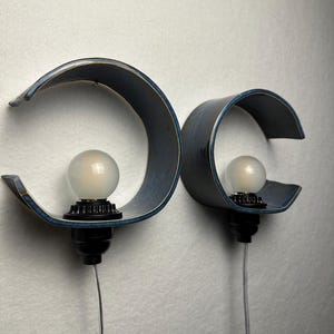 May include: Two wall-mounted lamps featuring curved, dark blue ceramic shades with a smooth finish. Each lamp includes a black socket base holding a round, frosted white light bulb. A thin, clear electrical cord extends downward from each fixture.