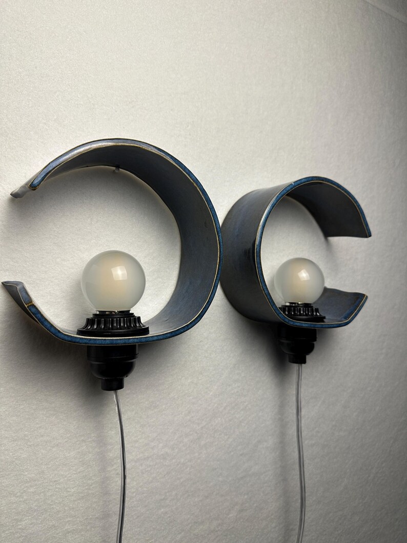 May include: Two wall-mounted lamps featuring curved, dark blue ceramic shades with a smooth finish. Each lamp includes a black socket base holding a round, frosted white light bulb. A thin, clear electrical cord extends downward from each fixture.