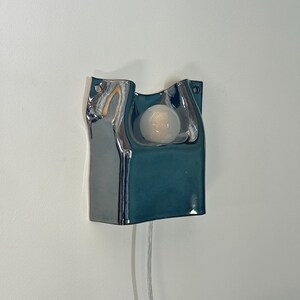 May include: A wall-mounted ceramic sconce featuring a glossy, deep teal finish with a fluid, organic shape. The fixture includes a central circular cutout holding a single frosted white light bulb and a thin, transparent power cord extending downward.
