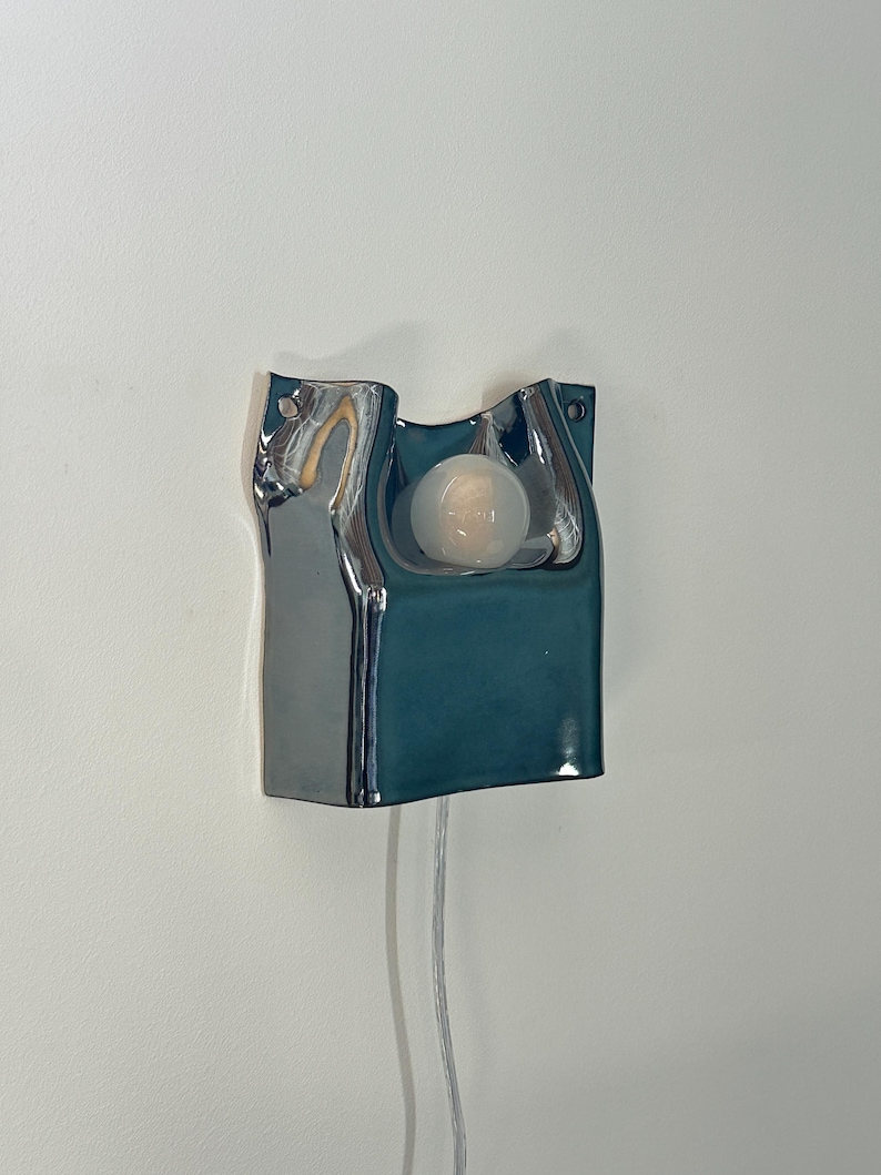 May include: A wall-mounted ceramic sconce featuring a glossy, deep teal finish with a fluid, organic shape. The fixture includes a central circular cutout holding a single frosted white light bulb and a thin, transparent power cord extending downward.
