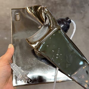Peut inclure: Une applique murale en c&eacute;ramique avec un vernis argent&eacute; m&eacute;tallique r&eacute;fl&eacute;chissant. La pi&egrave;ce pr&eacute;sente une base rectangulaire avec une extension sculpturale incurv&eacute;e, une douille noire pour ampoule et un cordon &eacute;lectrique transparent avec fiche.