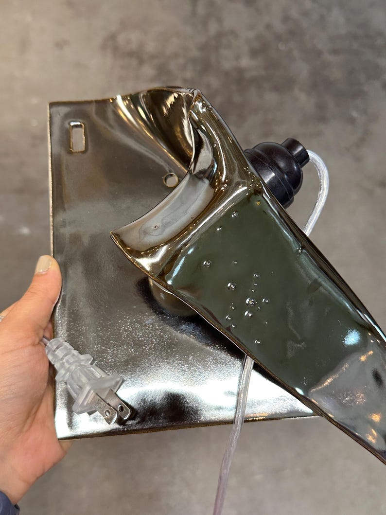 Peut inclure: Une applique murale en c&eacute;ramique avec un vernis argent&eacute; m&eacute;tallique r&eacute;fl&eacute;chissant. La pi&egrave;ce pr&eacute;sente une base rectangulaire avec une extension sculpturale incurv&eacute;e, une douille noire pour ampoule et un cordon &eacute;lectrique transparent avec fiche.