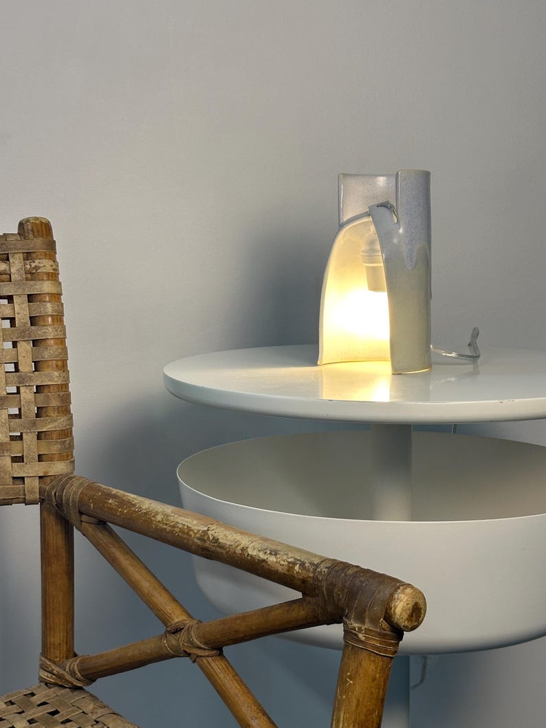 K&ouml;nnte beinhalten: Eine Keramik-Tischleuchte mit einem zylindrischen, hellblau und wei&szlig; glasierten K&ouml;rper, der eine geschwungene Aussparung aufweist. Die Lampe strahlt ein warmes, helles Licht von einer internen Gl&uuml;hbirne aus und hat ein d&uuml;nnes, transparentes Kabel.