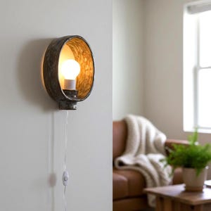 May include: A wall-mounted light fixture with a circular, metallic frame and a warm, golden interior. The fixture houses a lit bulb and is connected to a wall switch via a white cord. The design is modern and suitable for interior decor.