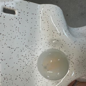 Może przedstawiać: Biały ceramiczny przedmiot z nakrapianym wzorem w małe ciemnobrązowe kropki. Powierzchnia ma gładkie, błyszczące wykończenie i zawiera okrągłe wgłębienie z p&oacute;łprzezroczystą, jasną substancją w środku oraz prostokątne wycięcie.