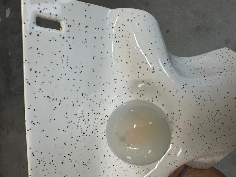 Może przedstawiać: Biały ceramiczny przedmiot z nakrapianym wzorem w małe ciemnobrązowe kropki. Powierzchnia ma gładkie, błyszczące wykończenie i zawiera okrągłe wgłębienie z p&oacute;łprzezroczystą, jasną substancją w środku oraz prostokątne wycięcie.