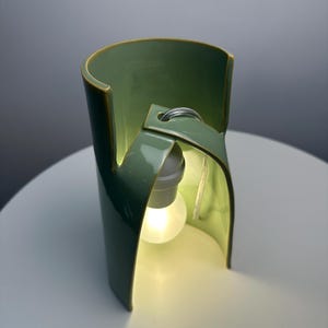 May include: A sage green ceramic table lamp with a curved, cylindrical structure that features an open front design. A single white light bulb is suspended from the top interior of the ceramic frame, emitting a soft, warm glow from within the piece.