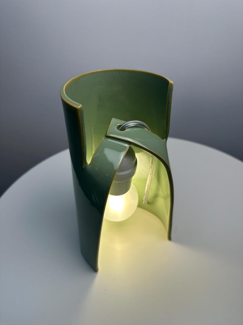 May include: A sage green ceramic table lamp with a curved, cylindrical structure that features an open front design. A single white light bulb is suspended from the top interior of the ceramic frame, emitting a soft, warm glow from within the piece.