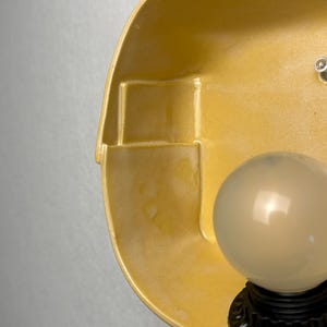 May include: A wall-mounted light fixture featuring a smooth, mustard yellow ceramic shade with a geometric, stepped interior design. A black base supports a single, spherical, frosted white glass light bulb that emits a soft, diffused glow.