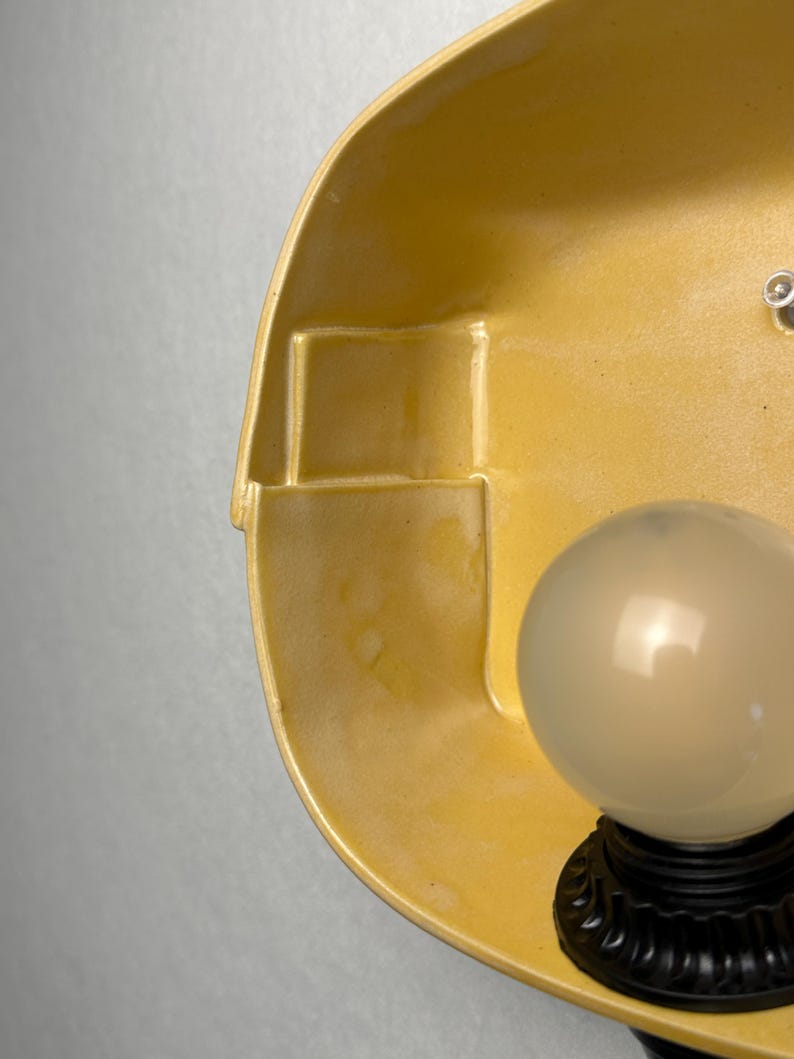 May include: A wall-mounted light fixture featuring a smooth, mustard yellow ceramic shade with a geometric, stepped interior design. A black base supports a single, spherical, frosted white glass light bulb that emits a soft, diffused glow.
