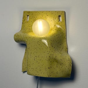 Puede incluir: Un aplique de pared de cer&aacute;mica con un esmalte amarillo brillante y motas negras. La forma es irregular y drapeada, con dos orificios de montaje rectangulares en la parte superior y una bombilla esf&eacute;rica brillante situada en el centro.