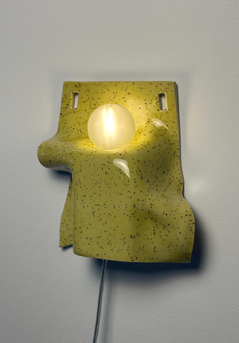 Puede incluir: Un aplique de pared de cer&aacute;mica con un esmalte amarillo brillante y motas negras. La forma es irregular y drapeada, con dos orificios de montaje rectangulares en la parte superior y una bombilla esf&eacute;rica brillante situada en el centro.
