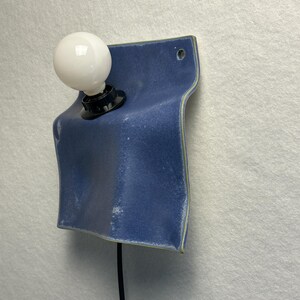 May include: A wall-mounted light fixture featuring a blue ceramic plate with a wavy, folded shape. A black socket holds a round white light bulb on the left side. A black power cord extends downward from the base of the socket.