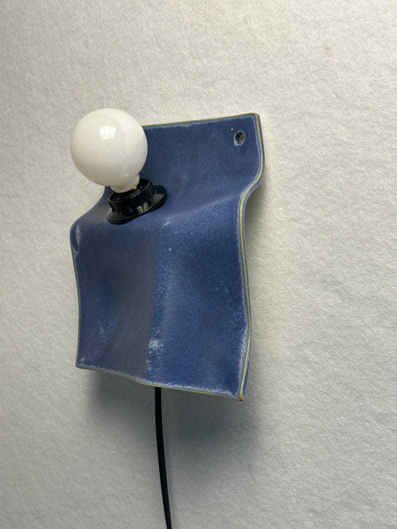 May include: A wall-mounted light fixture featuring a blue ceramic plate with a wavy, folded shape. A black socket holds a round white light bulb on the left side. A black power cord extends downward from the base of the socket.