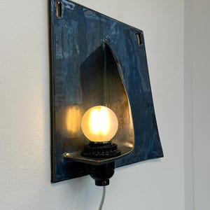 May include: A wall-mounted light fixture with a square, blue-toned metal shade. The fixture has a curved opening to house a black light socket and a round, frosted light bulb. A gray cord extends from the base.