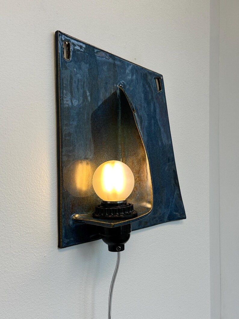 May include: A wall-mounted light fixture with a square, blue-toned metal shade. The fixture has a curved opening to house a black light socket and a round, frosted light bulb. A gray cord extends from the base.