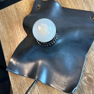 May include: A wall-mounted lamp featuring a matte black, irregularly shaped, wavy metal plate with two small circular mounting holes at the top corners. A single round, white frosted glass light bulb is centered on the plate in a black socket.