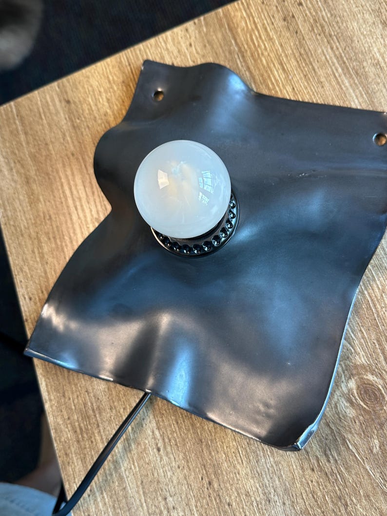 May include: A wall-mounted lamp featuring a matte black, irregularly shaped, wavy metal plate with two small circular mounting holes at the top corners. A single round, white frosted glass light bulb is centered on the plate in a black socket.