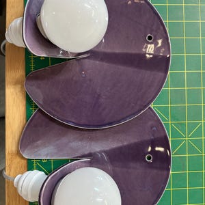 K&ouml;nnte beinhalten: Zwei violette Wandleuchten aus Keramik, jede mit einem kreisf&ouml;rmigen, leicht gew&ouml;lbten Plattendesign und einem Ausschnitt. Eine wei&szlig;e, kugelf&ouml;rmige Gl&uuml;hbirne ist an der Seite jeder Leuchte befestigt, und ein kleines Loch befindet sich rechts.