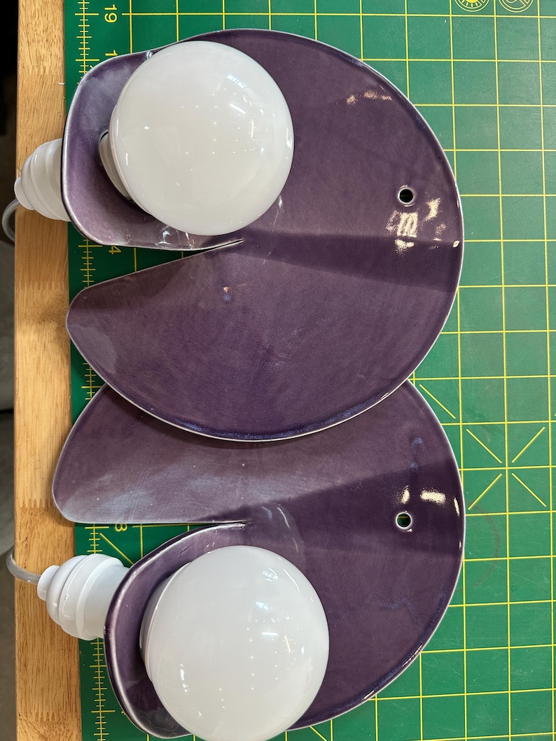 K&ouml;nnte beinhalten: Zwei violette Wandleuchten aus Keramik, jede mit einem kreisf&ouml;rmigen, leicht gew&ouml;lbten Plattendesign und einem Ausschnitt. Eine wei&szlig;e, kugelf&ouml;rmige Gl&uuml;hbirne ist an der Seite jeder Leuchte befestigt, und ein kleines Loch befindet sich rechts.