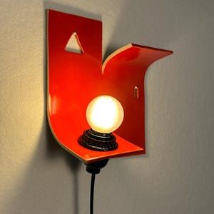 May include: A contemporary wall light in a vibrant red hue, with a curved shape and a small shelf. It holds a spherical, illuminated light bulb with a black base. The design includes a triangular cut-out and a black power cable.