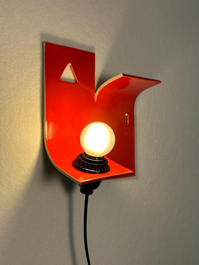 May include: A contemporary wall light in a vibrant red hue, with a curved shape and a small shelf. It holds a spherical, illuminated light bulb with a black base. The design includes a triangular cut-out and a black power cable.