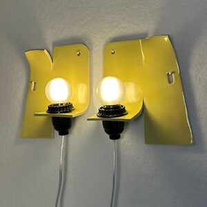 May include: A pair of wall-mounted sconces featuring bright yellow, irregularly shaped metal backplates. Each fixture includes a black socket holding a glowing, spherical white light bulb. A thin, clear power cord extends downward from each lamp.