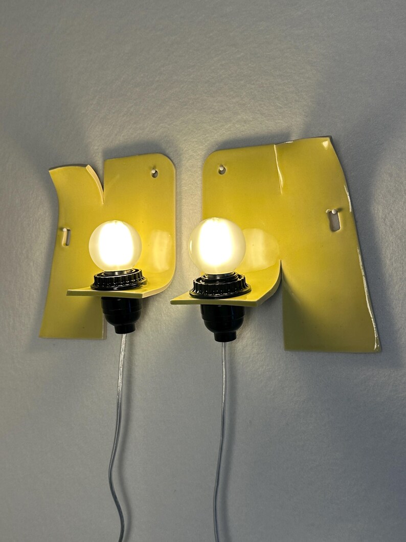 May include: A pair of wall-mounted sconces featuring bright yellow, irregularly shaped metal backplates. Each fixture includes a black socket holding a glowing, spherical white light bulb. A thin, clear power cord extends downward from each lamp.