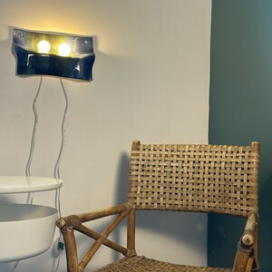 May include: A wall-mounted light fixture with a curved, dark blue and yellow glass shade featuring two glowing bulbs, positioned above a woven rattan chair with a cross-brace frame and a white tiered side table with a visible power cord.