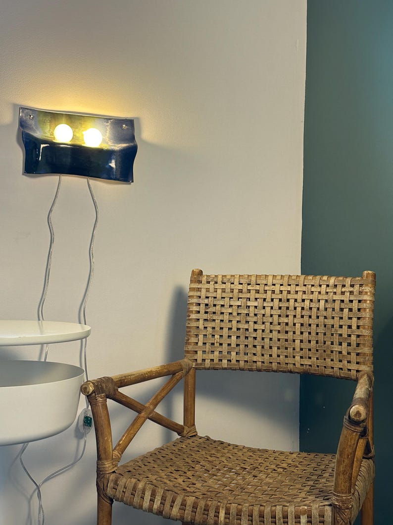May include: A wall-mounted light fixture with a curved, dark blue and yellow glass shade featuring two glowing bulbs, positioned above a woven rattan chair with a cross-brace frame and a white tiered side table with a visible power cord.