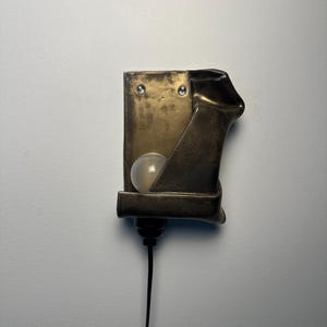 May include: A wall-mounted lamp featuring a rectangular, metallic bronze-toned fixture with a folded, sculptural edge. A single round, frosted white light bulb is nestled within the structure, and a black power cord extends from the bottom base.