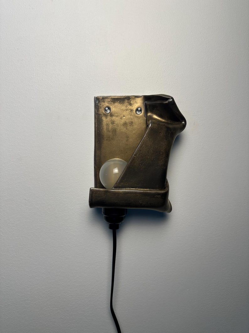 May include: A wall-mounted lamp featuring a rectangular, metallic bronze-toned fixture with a folded, sculptural edge. A single round, frosted white light bulb is nestled within the structure, and a black power cord extends from the bottom base.