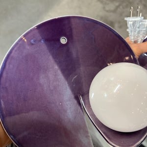 K&ouml;nnte beinhalten: Eine kreisf&ouml;rmige Wandleuchte aus violetter Keramik mit gl&auml;nzender Oberfl&auml;che und einem kleinen runden Loch am oberen Rand. Die Leuchte verf&uuml;gt &uuml;ber eine wei&szlig;e, kugelf&ouml;rmige Gl&uuml;hbirne an der Seite und ein transparentes Stromkabel.