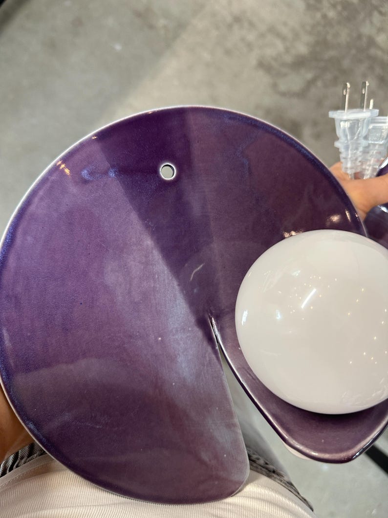 K&ouml;nnte beinhalten: Eine kreisf&ouml;rmige Wandleuchte aus violetter Keramik mit gl&auml;nzender Oberfl&auml;che und einem kleinen runden Loch am oberen Rand. Die Leuchte verf&uuml;gt &uuml;ber eine wei&szlig;e, kugelf&ouml;rmige Gl&uuml;hbirne an der Seite und ein transparentes Stromkabel.