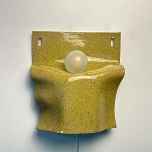 May include: A wall-mounted sconce featuring a glossy, speckled chartreuse ceramic shade with a wavy, organic form. A single, spherical, frosted white glass light bulb is centered in the middle of the fixture, which includes two mounting holes.