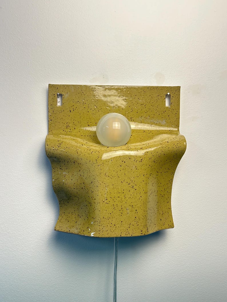 May include: A wall-mounted sconce featuring a glossy, speckled chartreuse ceramic shade with a wavy, organic form. A single, spherical, frosted white glass light bulb is centered in the middle of the fixture, which includes two mounting holes.