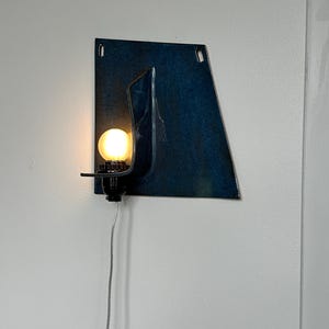 May include: A modern wall sconce with a dark blue, abstract metal design. The sconce features a small, round light bulb emitting a warm glow. A black fixture holds the bulb, and a silver cord extends downward.