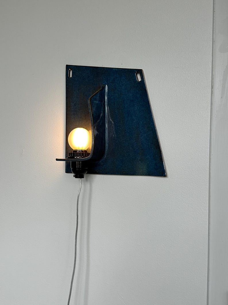 May include: A modern wall sconce with a dark blue, abstract metal design. The sconce features a small, round light bulb emitting a warm glow. A black fixture holds the bulb, and a silver cord extends downward.