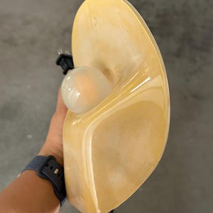 May include: A wall-mounted lamp featuring a smooth, curved, amber-colored resin shade with a glossy finish. The lamp includes a round, frosted white light bulb centered within the shade and a black electrical cord with a standard plug attached.