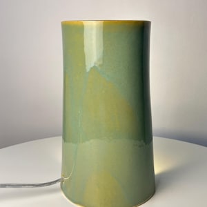 May include: A cylindrical ceramic table lamp with a smooth, glossy glaze in shades of sage green and mustard yellow. The lamp features a small circular opening at the base for a thin, clear electrical cord to pass through, emitting a soft light.