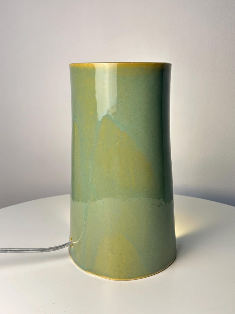 May include: A cylindrical ceramic table lamp with a smooth, glossy glaze in shades of sage green and mustard yellow. The lamp features a small circular opening at the base for a thin, clear electrical cord to pass through, emitting a soft light.