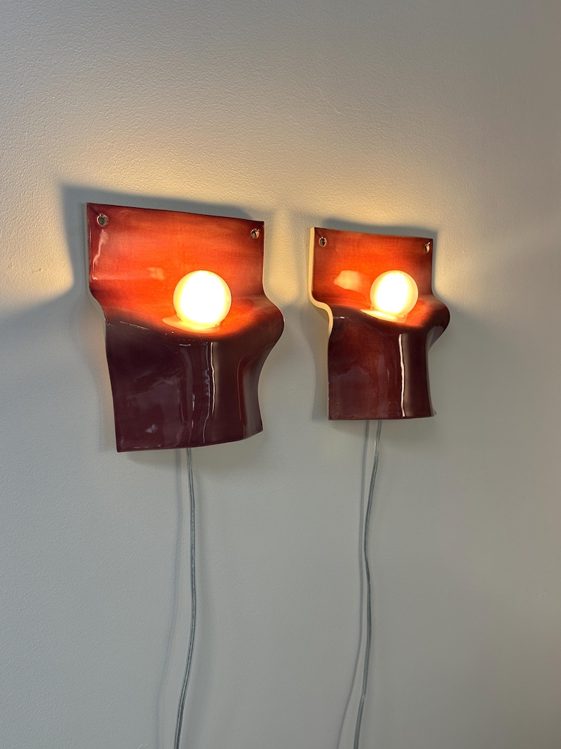 May include: A pair of wall-mounted sconces featuring curved, glossy red ceramic shades with a gradient finish. Each fixture holds a single glowing spherical light bulb and includes a thin, light-colored electrical cord extending downward.