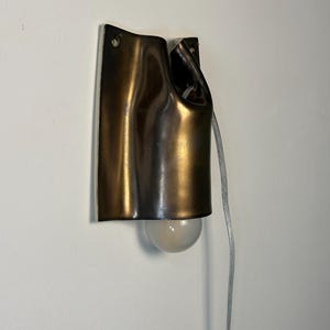 May include: A wall-mounted lamp featuring a dark bronze-toned, curved metal shade with a fluid, draped appearance. The fixture includes a single exposed round light bulb at the bottom and a clear electrical cord extending from the side.