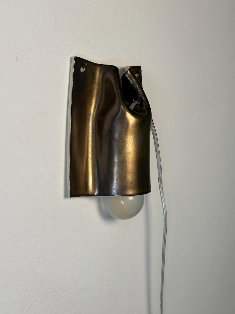 May include: A wall-mounted lamp featuring a dark bronze-toned, curved metal shade with a fluid, draped appearance. The fixture includes a single exposed round light bulb at the bottom and a clear electrical cord extending from the side.